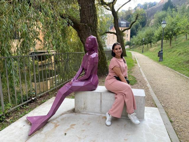 BellaBrownX sits on a stone bench, wearing pink pants and white sneakers, next to a purple geometric sculpture, with a tree and grassy path in the background.