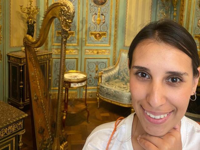 BellaBrownX with brown eyes, dark hair, white shirt, smiling, hand on chin, gold harp, ornate blue-green wall, wooden floor, vintage furniture.