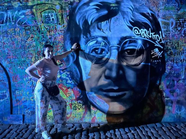 BellaBrownX stands with hand on graffiti wall featuring a blue, oversized face with round glasses. She's wearing a sleeveless top and patterned pants, against cobblestone.