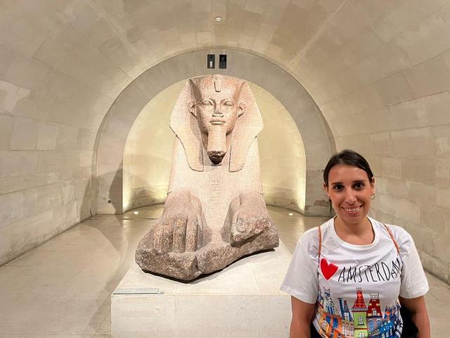 BellaBrownX with small tits, wearing a white shirt with colorful graphics, stands in front of a large, ancient Egyptian statue of a seated deity with a calm face.
