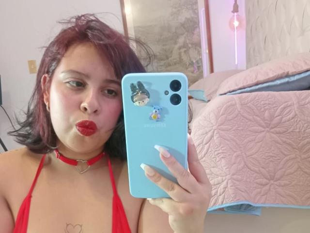 RouxDeVille with red hair, red choker, and red bikini top, pouting with red lipstick, holding a blue phone with stickers, in a bedroom with a quilted bed.