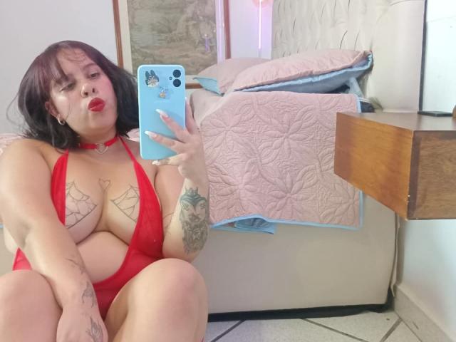 RouxDeVille in a red halter bikini, puffy lips, dark bob, tattoos on chest and arm, taking a mirror selfie, showing her big tits, pink quilted bed in background.