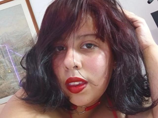 RouxDeVille with dark, wavy hair, red lipstick, and a red choker with a heart. She's touching her head, showing a slightly flushed face and soft, brown eyes.