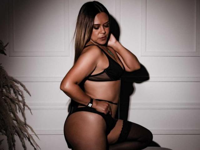 LilithBonneth in black lingerie, showing her tits and ass, with long brown hair, a wristwatch, and a sexy side profile, standing against a white wall.