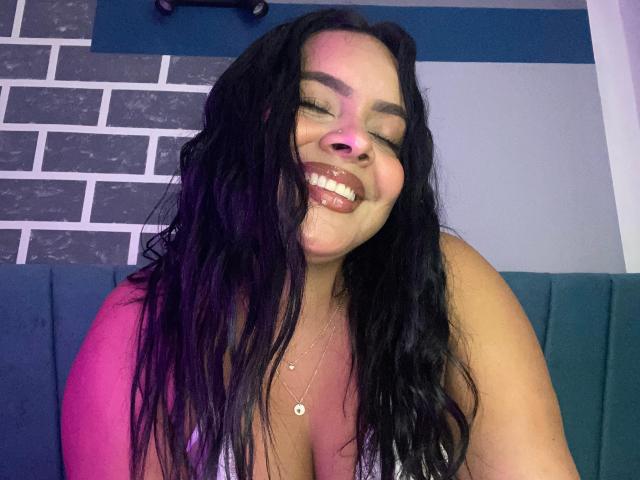 Amaranta Carey ha i capelli neri e mossi, un viso rotondetto e un grande sorriso che mostra i denti. Indossa una maglietta rosa, e il suo seno è visibile. È seduta su un divano blu..