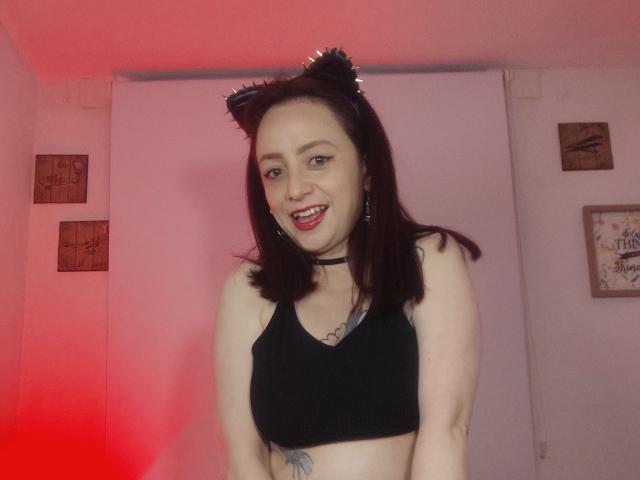 CatalinaMonyque with dark cat ears, black choker, red lipstick, black sports bra, showing her tits, and a tattoo on her side, smiling in a red-lit room.