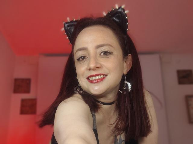 CatalinaMonyque con rossetto rosso, orecchie da gatto nere e orecchini a forma di mezzaluna, sorride alla fotocamera, mostrando una piccola goccia di liquido bianco sul mento..