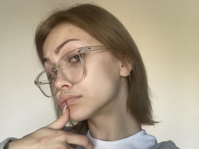LolaMi ha i capelli castano chiaro, occhiali trasparenti e un'espressione pensierosa, con un dito che tocca il labbro inferiore. Indossa una maglietta grigia e bianca..