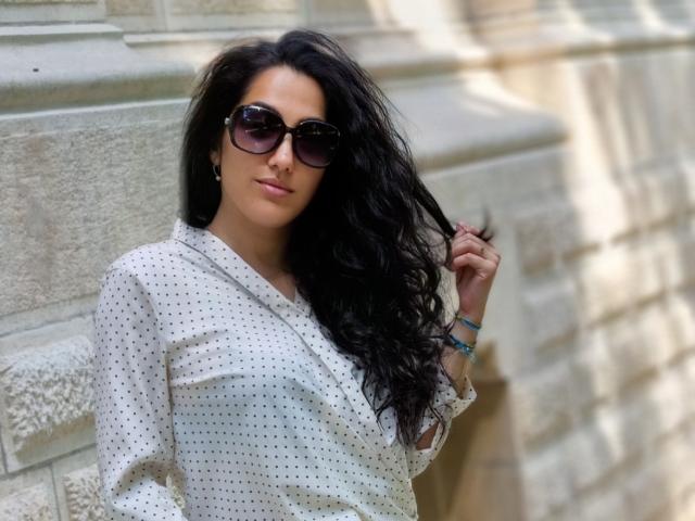 SexyMirra, avec de longs cheveux noirs, portant une chemise blanche à pois, des lunettes de soleil noires et un sourire discret, tout en touchant ses cheveux. Elle se tient contre un mur de pierre..