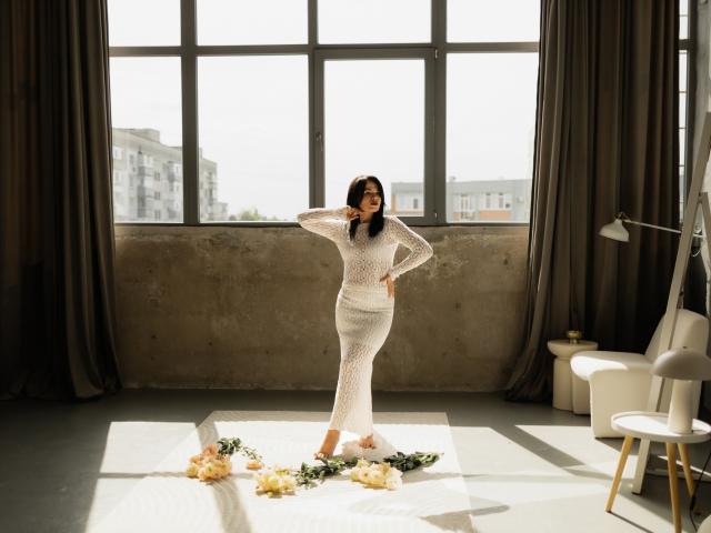 JasmineKouch stands in a sunlit room wearing a white, long-sleeve, see-through dress, with her hand on her hip, and a floral arrangement at her feet.
