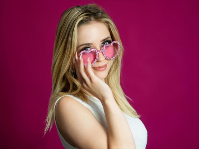 ClaudiaNice with blonde hair, wearing pink heart-shaped glasses with sparkles, white sleeveless top, smiling, hand gently touching glasses, pink background.