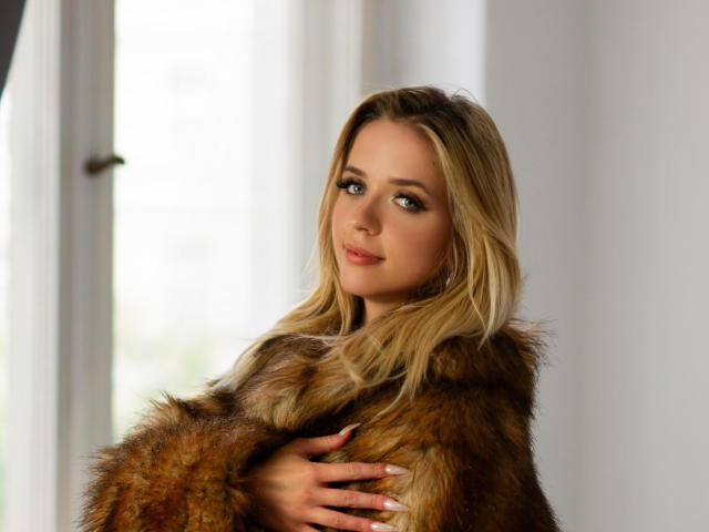 ClaudiaNice with blonde hair, wearing a brown fur coat, holding it with her hand, looking at the camera with a slight smile, soft natural light.