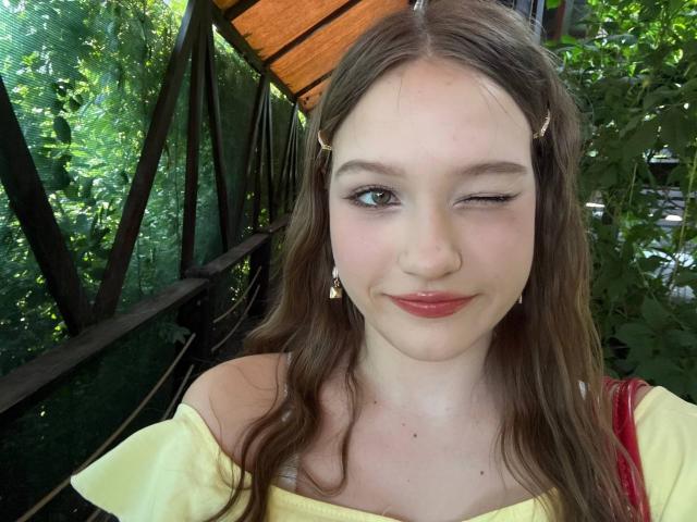 Bubochka with winking eye, pink lips, and yellow dress, has brown hair, small earrings, and a cute smile. Her tits are not visible. She's standing outside in greenery.