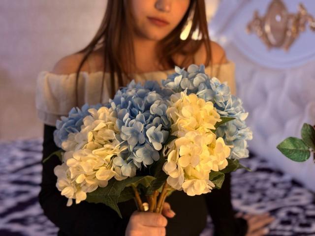Bubochka sostiene un ramo de hortensias azules y blancas, lleva una blusa negra y blanca con un hombro descubierto, tiene el cabello castaño y la luz es suave y cálida..