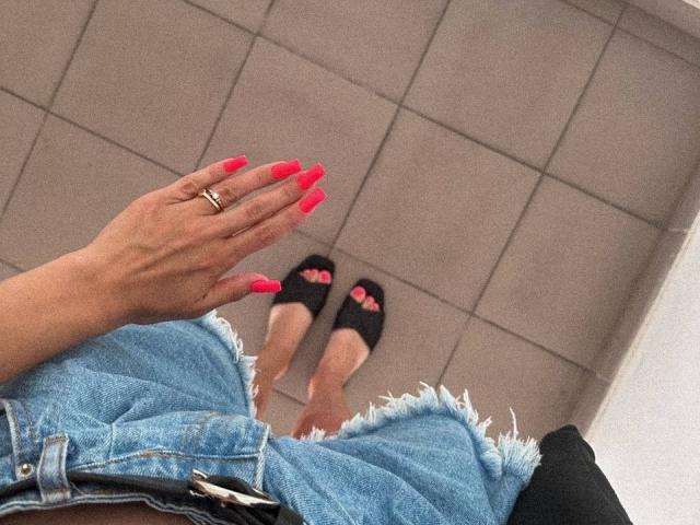 Siyana's hand with bright pink nails and ring, wearing frayed denim shorts, black flats with pink toenails, visible from a downward angle.