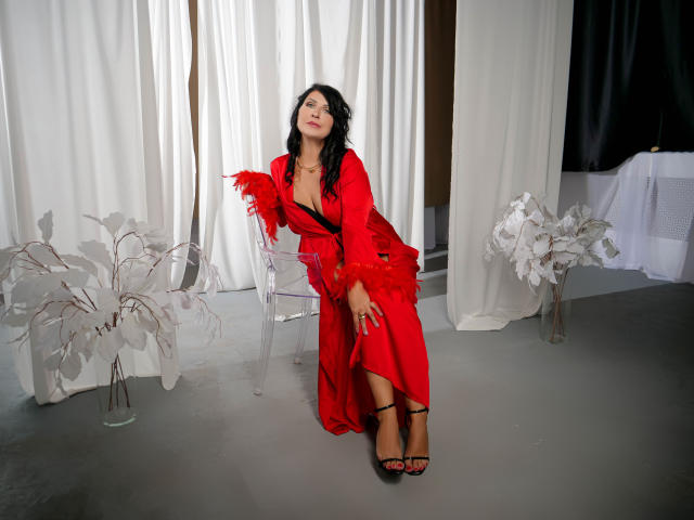 MoonVelour with black hair, red satin robe, and black high heels, leaning on a chair, showing her tits in a sexy pose, standing in a grey room.