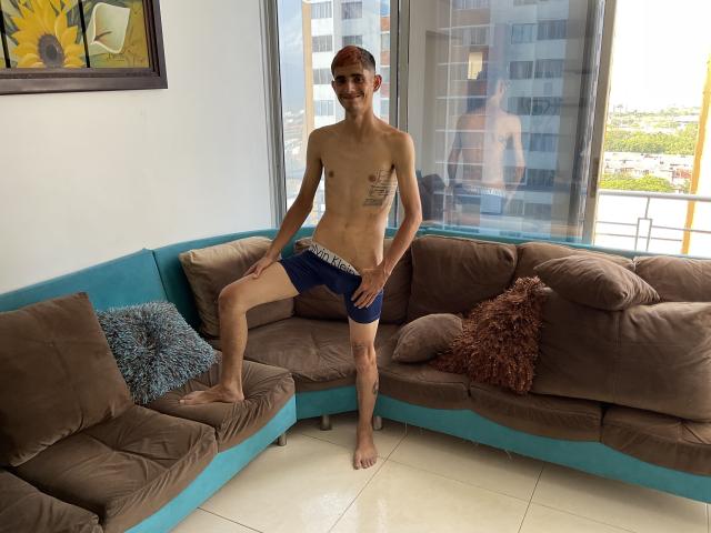 SkinnyAngie stands on a brown and blue sofa, wearing blue boxer shorts, with one leg up, showing a tattoo on the left side of his chest. Barefoot, smiling.