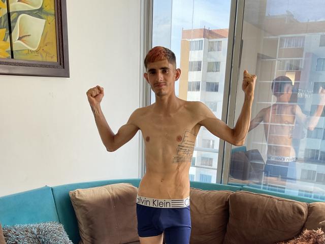 SkinnyAngie stands in blue underwear, fists raised, showing a slim, tattooed torso with red hair, reflecting in a window. She's in a room with a couch and art.