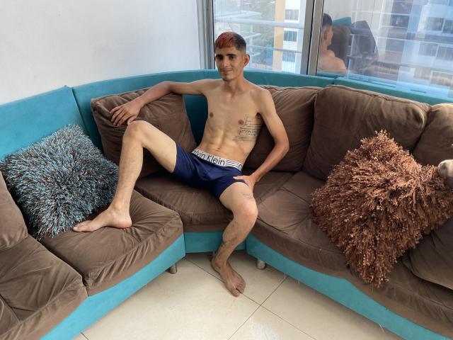 SkinnyAngie, shirtless with a slim, toned body and small tattoos on the side, sits on a brown and blue couch wearing dark blue underwear, legs spread, with a fluffy pillow.