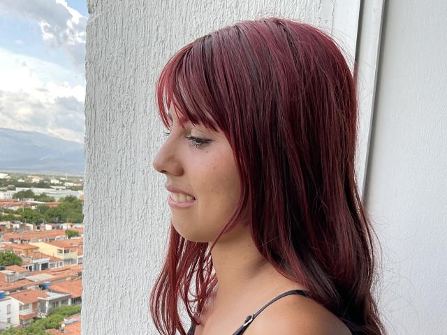 SkinnyAngie with red and black hair, smiling sideways, showing her nose and small tits, wearing a black strap, standing by a white textured wall.