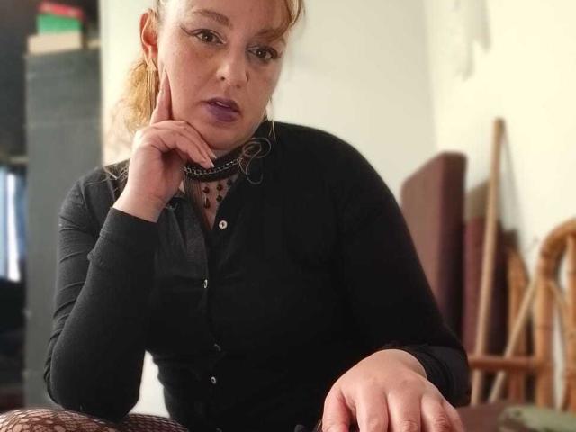 AmandaHotL with pale skin, red hair in a ponytail, wearing a black long-sleeve shirt, purple lipstick, and a black choker, resting her chin on her hand.
