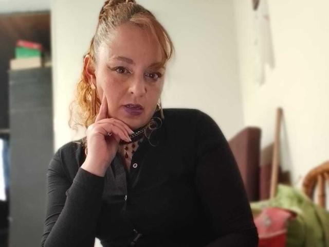 AmandaHotL with light brown hair in a ponytail, black long-sleeve shirt, purple lipstick, and hoop earrings, resting her chin on her hand, looking serious.