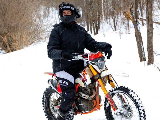 MisterHardcore montando una motocicleta roja y blanca sucia, usando un casco, una máscara, una chaqueta y guantes negros, con pantalones y botas negros, en un bosque nevado..