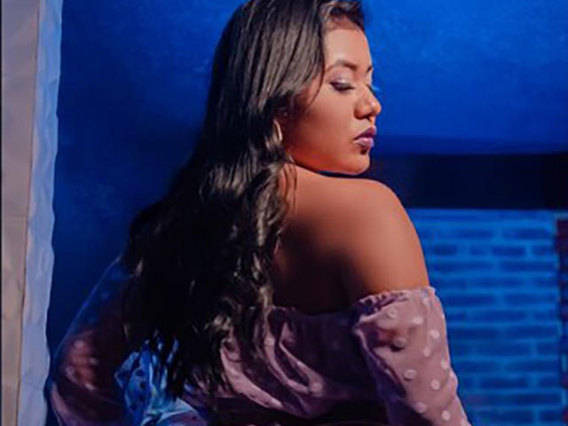LiaDecker with long, wavy brown hair, wearing an off-shoulder purple polka-dot top, looking over her shoulder with a slight pout, under blue lighting.