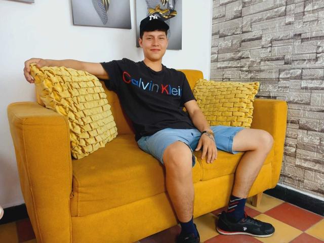 DanielOn, com uma estrutura física esguia, veste uma camisa preta da Calvin Klein, shorts azuis e tênis pretos. Ele está sentado em um sofá amarelo, com os braços relaxados e as pernas abertas, e travesseiros amarelos com babados atrás dele..