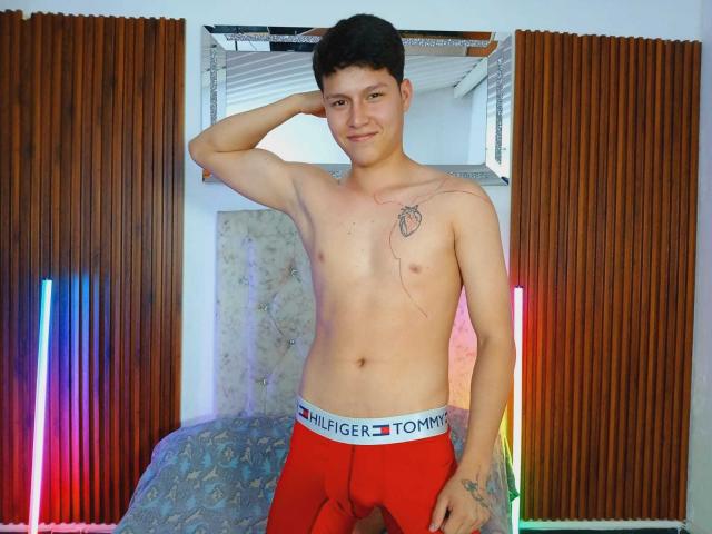 Daniel sin camisa, con cabello negro corto, ropa interior roja Tommy, un tatuaje pequeño en el pecho, de pie con un brazo detrás de la cabeza, sonriendo, luces fluorescentes a los lados, un tatuaje en la muñeca izquierda..