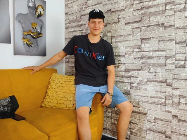 DanielOn es un joven con una camiseta negra de Calvin Klein, pantalones cortos azules y una gorra negra, sentado en un sofá amarillo, mostrando su trasero, con una sonrisa relajada..