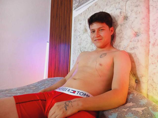 DanielOn, sin camisa y con calzoncillos rojos, está sentado en una cama, sonriendo, con tatuajes en el pecho y la mano, cabello castaño corto, piel clara, con un poco de vello en el pecho, apoyado contra una pared con estampado floral..