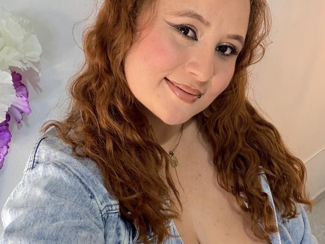IsabellaHotest with wavy red hair, light skin, and black eyeliner, smiling with a nose ring, wearing a denim shirt, showing cleavage, and a necklace, in a selfie.