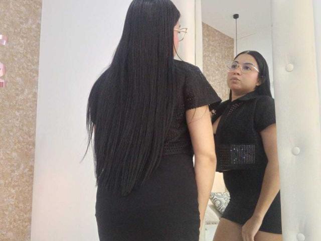 AliceSmity with long black hair, glasses, black mesh top, and black skirt, standing in front of mirror, looking at reflection, showing her ass and small tits.