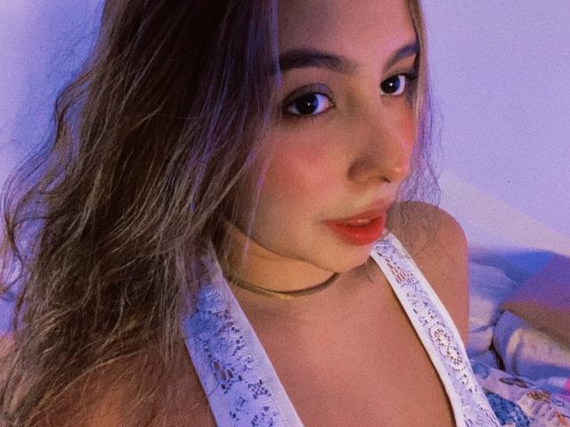 NicoleSweetyPie with wavy brown hair, wearing a white lace halter top, has her lips slightly parted, showing her red lipstick, and a gold choker around her neck.