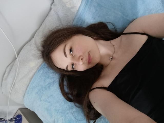 LilithDiva lying on a bed, wearing a black camisole, with long brown hair, light skin, and a small necklace, looking at the camera, with a blue pillow behind her.