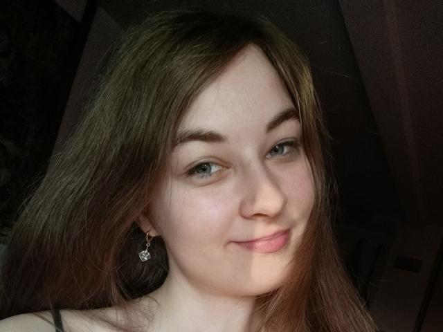 LilithDiva with light skin, blue eyes, and brown hair, smiling slightly, wearing small diamond earrings, in a dimly lit room.