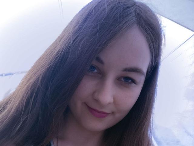 LilithDiva has long brown hair, blue eyes, and a slight smile with pink lipstick, looking cute and innocent. Her small tits are barely visible in the shot.