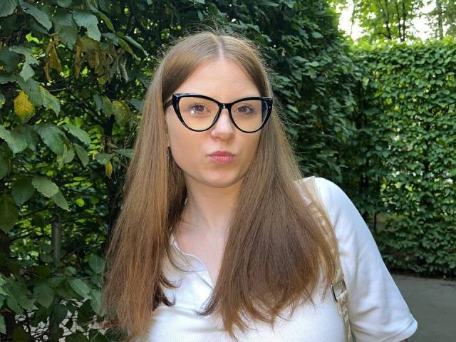 FoxyTeaser has straight brown hair, black glasses, and a white shirt, puckering her lips in a playful manner. Her ass is not visible. She's surrounded by green foliage.
