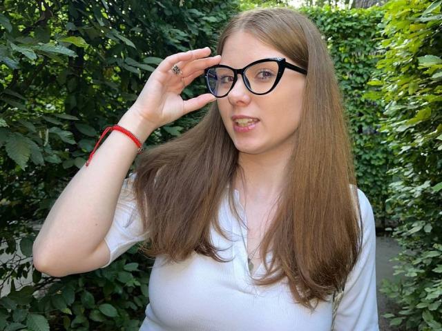 FoxyTeaser with long brown hair, black glasses, white shirt, and red bracelet, adjusting glasses, showing her tits slightly through her shirt, standing in lush green foliage.