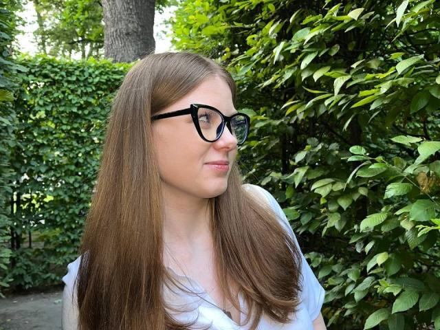 FoxyTeaser has long brown hair, black glasses, and a slight smile, wearing a white shirt, with green leafy bushes in the background. Her tits are small and cute.