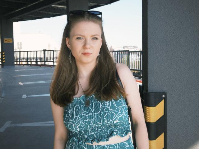 FoxyTeaser has long brown hair, blue eyes, and wears a teal patterned top with ruffles, standing in a parking garage, sunglasses on head, black and yellow bollard beside her.