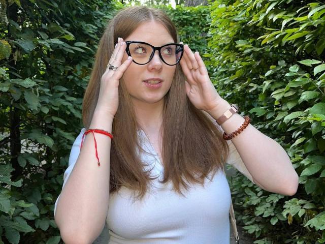 FoxyTeaser has brown hair, wearing black glasses, white shirt, and red bracelet, adjusting glasses with hands, looking slightly to the side, green leafy background.