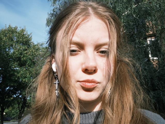 FoxyTeaser has light brown hair in pigtails, wearing cross earrings, with a serious expression, in bright sunlight, showing pale skin and slightly puffy lips.
