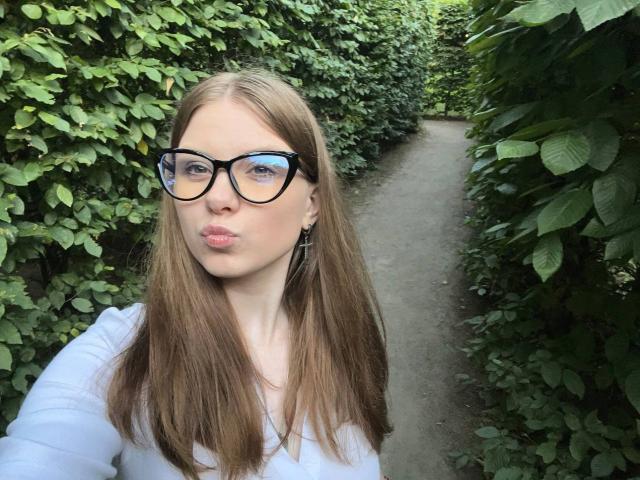 FoxyTeaser with pouty lips, wearing black glasses, white top, and brown straight hair, standing in a green leafy path. Boobs not visible, ass not visible, pussy not visible.