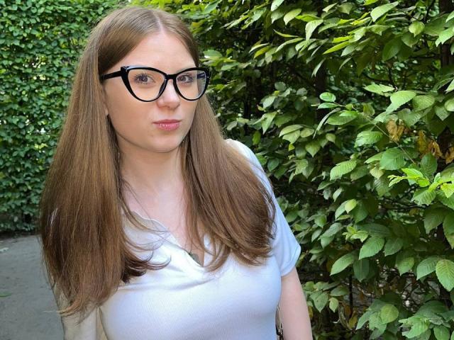 FoxyTeaser with long brown hair, black glasses, white shirt, and pouty lips, standing in front of green leafy bushes, showing small tits.