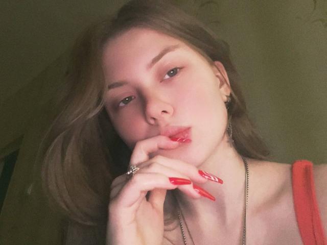 FoxyTeaser with pale skin, brown hair, red lipstick, red nails, finger touching lips, wearing red top, necklace, and earrings, looking seductive with subtle eye makeup.