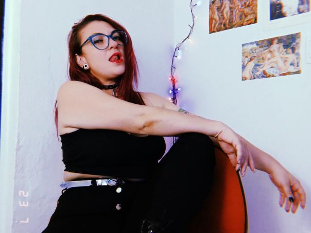 LunaRausch with red hair, glasses, black top, black pants, choker, sitting on orange chair, arms resting, mouth slightly open, wall with pictures and string lights behind.