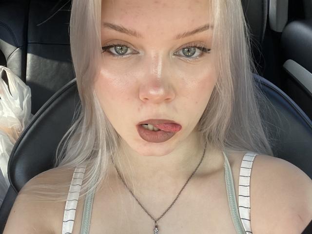 KaroasLove has pale skin, platinum blonde hair, green eyes, and is sticking her tongue out. She's wearing a white tank top with striped straps, and a necklace.