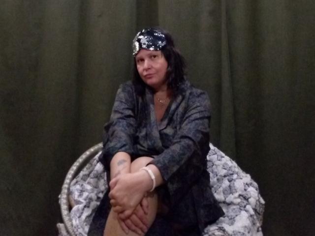 MiraaSweet, sitting with knees up, wearing a dark grey coat, black skirt, and a black floral headscarf, has a tattoo on her left forearm and a bracelet on her right.